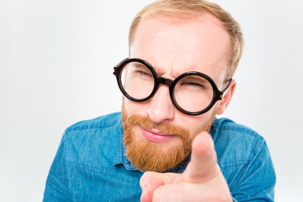 Portrait of suspicious young bearded man in funny round glasses pointing on you isolated over white background