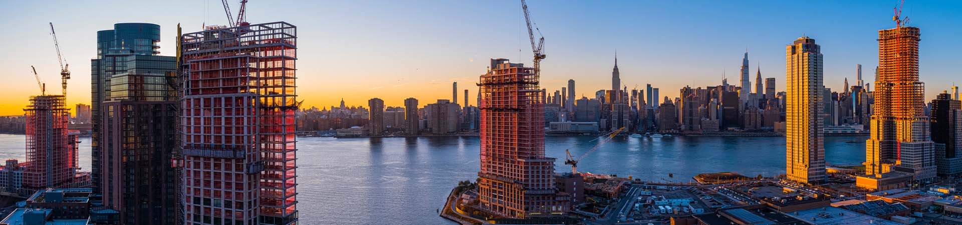 tall buildings and construction cranes beside a city river at sunset